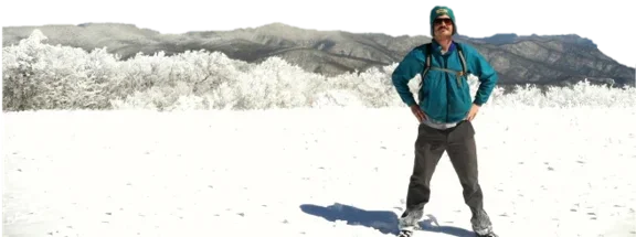 Stephen in a teal jacket, hands on his hips, standing in fresh snow with frost-covered mountains behind him.