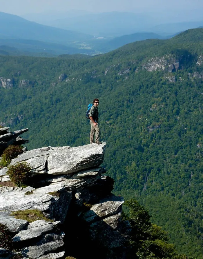 Stephen in a ranger uniform and blue pack, standing on a rock ledge high above a green forested gorge.