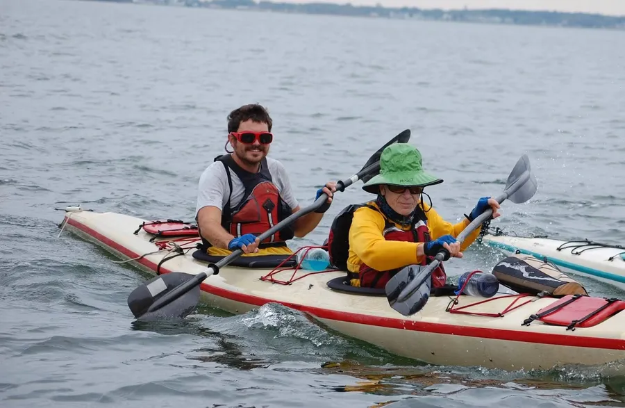 Stephen in red sunglasses and a life vest, smiling from the front seat of a two-person sea kayak with a paddler in a green sun hat behind him.