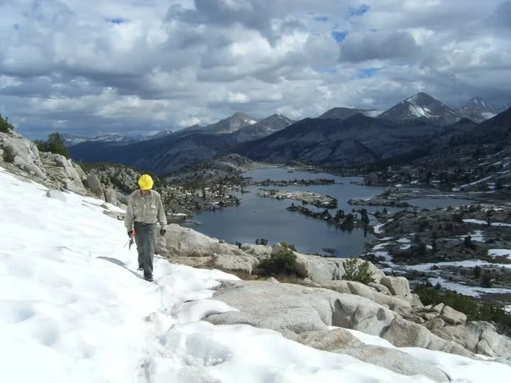 Stephen in a yellow hard hat walking across a snowfield above an alpine lake and mountains.