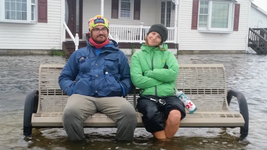 Stephen in a blue jacket and bright knit hat sits on a bench beside his wife in a green jacket, both with their feet in flood water in front of a white house.