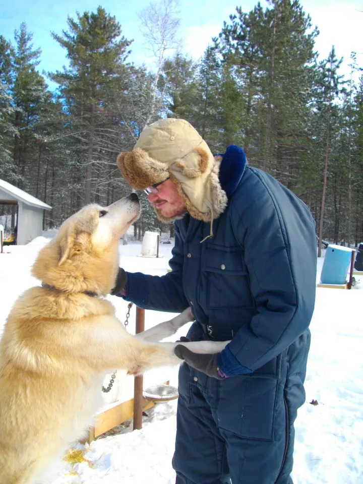 Stephen in a fur-lined winter hat and a blue insulated coat, nose to nose with Luke, a tall cream-colored sled dog standing on his hind legs in the snow.